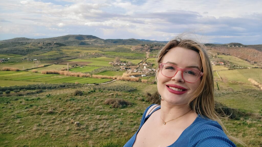 A blonde woman with dark pink lipstick and pink eyeglasses standing in front of peaceful agricultural green fields in Albania