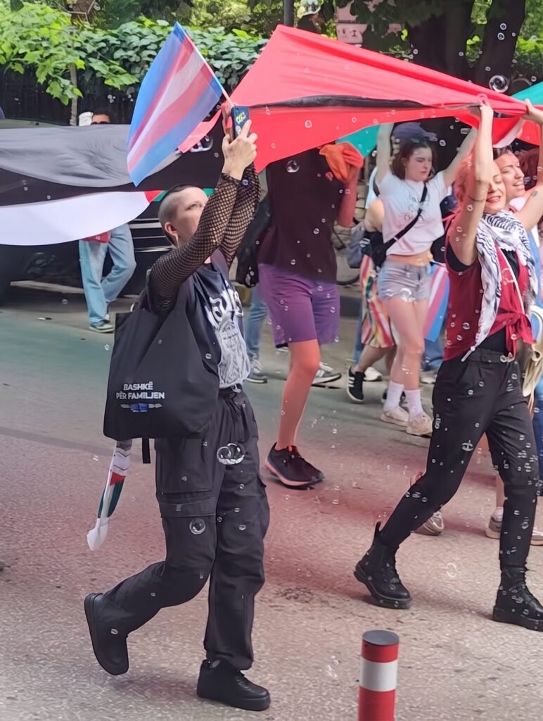 A sharp photograph from within a Pride parade on a street lined with trees. The central figure, with a buzzcut and black mesh top, holds up a trans pride flag (light blue, pink, white) while walking. They carry a tote bag with text that appears to be "BASHKË PËR FAMILJEN" (Together for Family). Behind them, a large red flag is held aloft, and floating bubbles catch the light.