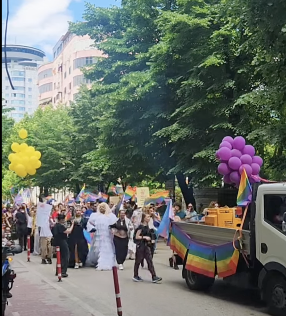 A colorful pride parade with drag queens, rainbow flags, and marchers in Tirana Albania with large trees on the side of the street