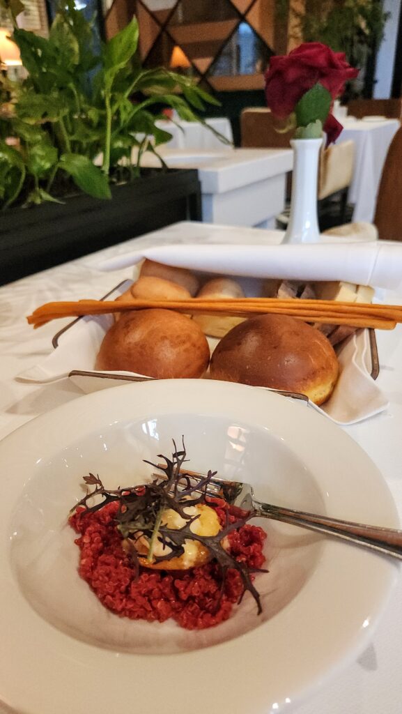 Plate with beetroot-colored couscous or grains and garnish, accompanied by a bread basket and white napkin on a table; a red rose in a vase is in the background.