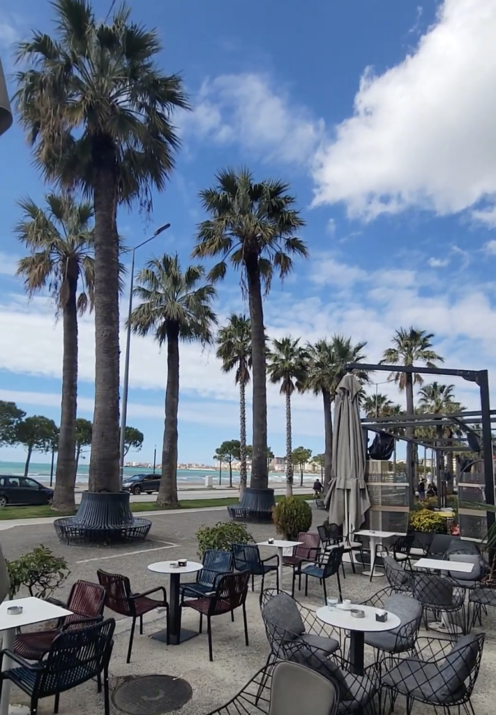 Outdoor cafe seating area under tall palm trees along the sunny Vlorë promenade in Albania.