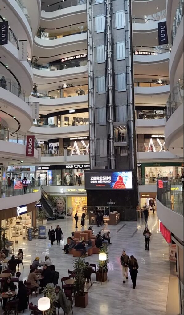 The modern, multi-level central atrium of Toptani Shopping Center in Tirana, Albania, showing glass elevators, curved white balconies, retail storefronts like Armani Exchange and Hugo, and a large digital sale advertisement.