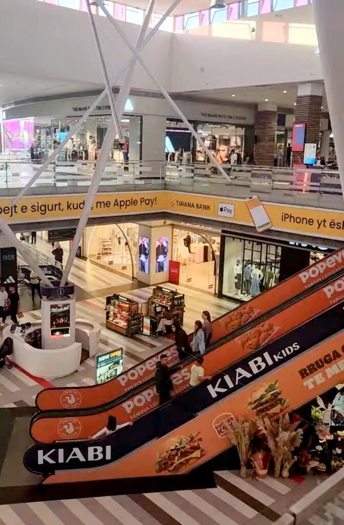 The multi-level interior of the Tirana East Gate (TEG) shopping mall in Albania, featuring escalators with advertisements for Popeyes and local retail shops like Kiabi.