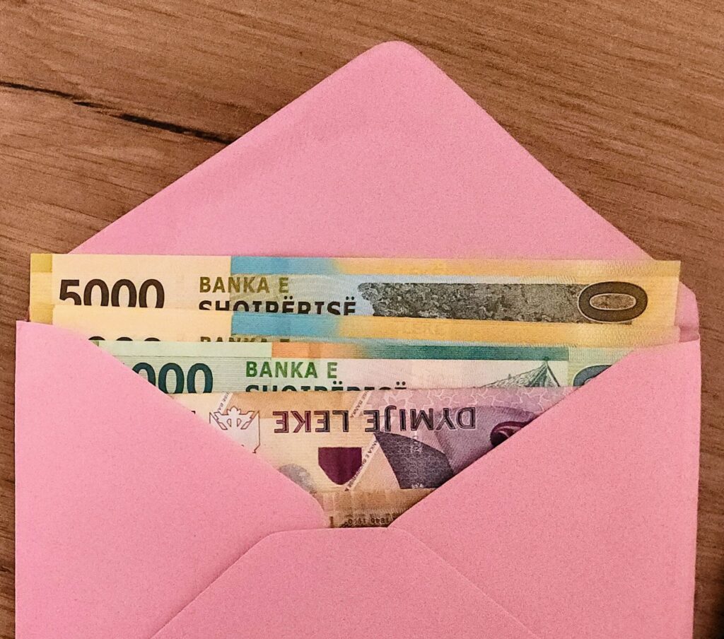 Several Albanian Lek banknotes, including 5000 and 2000 Lek bills from Banka e Shqipërisë, peeking out of a bright pink paper envelope on a wooden surface